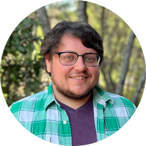 A headshot of an adult man with a beard and glasses. He is wearing a green and white checkered shirt over a purple shirt.