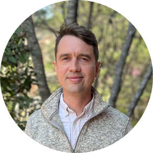 A headshot of an adult man with short brown hair. He is wearing a white button-down under a gray pullover.
